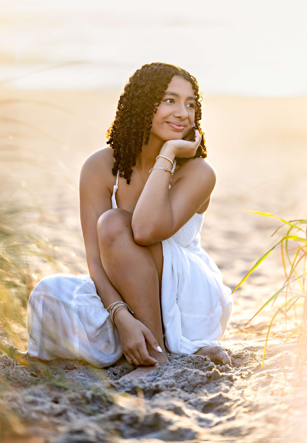 Senior photo on Cocoa Beach with a girl in a white dress looking off to the side, surrounded by beach grass and sand.