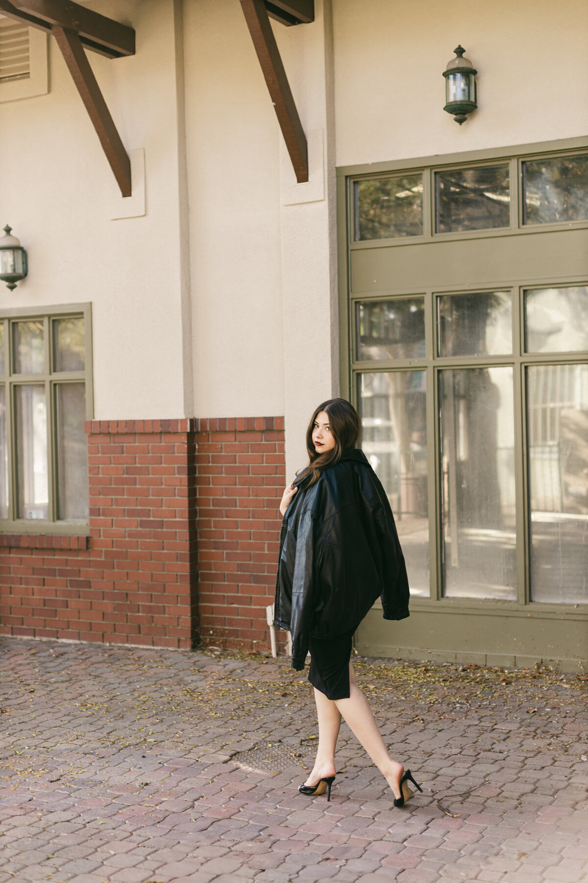 Stylish editorial branding photo of a woman in a black outfit and heels walking past an urban brick-and-stucco building — modern, confident, and fashion-forward.