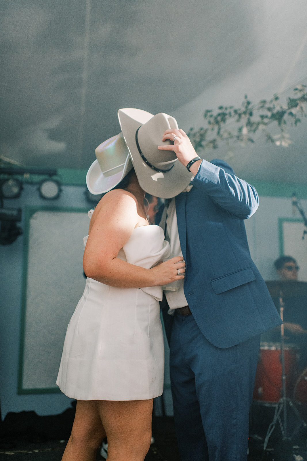  Bride and groom share a kiss behind cowboy hats during their late-night outfit change at a mountain wedding reception.