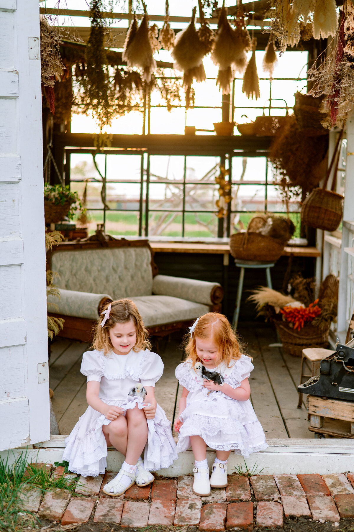 Mother with two young daughters sitting on a vintage sofa surrounded by florals and sunlight, photographed in Frisco, Texas by Jennifer L. Kirk Photography.