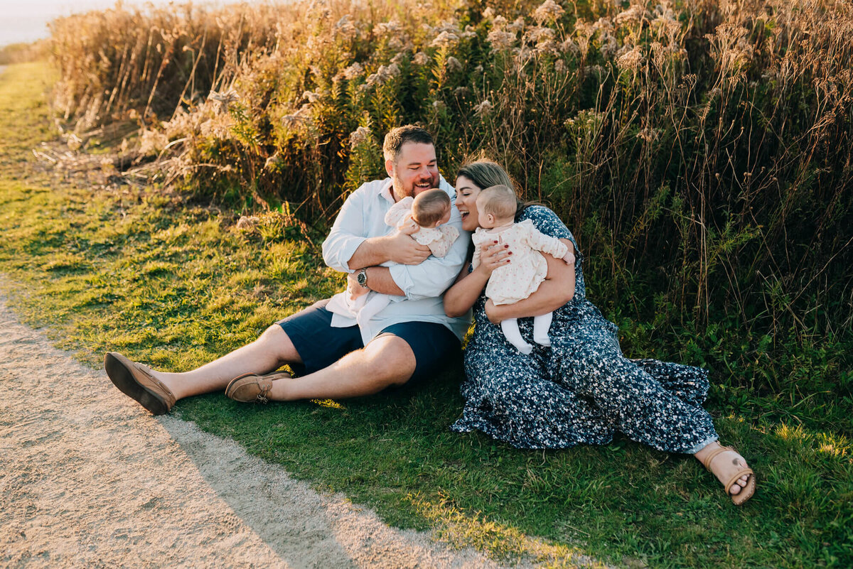A mother and father sit on the ground near the beach at sunset with their twin baby girls