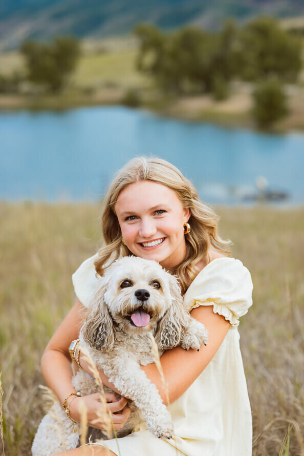 Senior girl snuggles her white dog with a blue lake behind her.