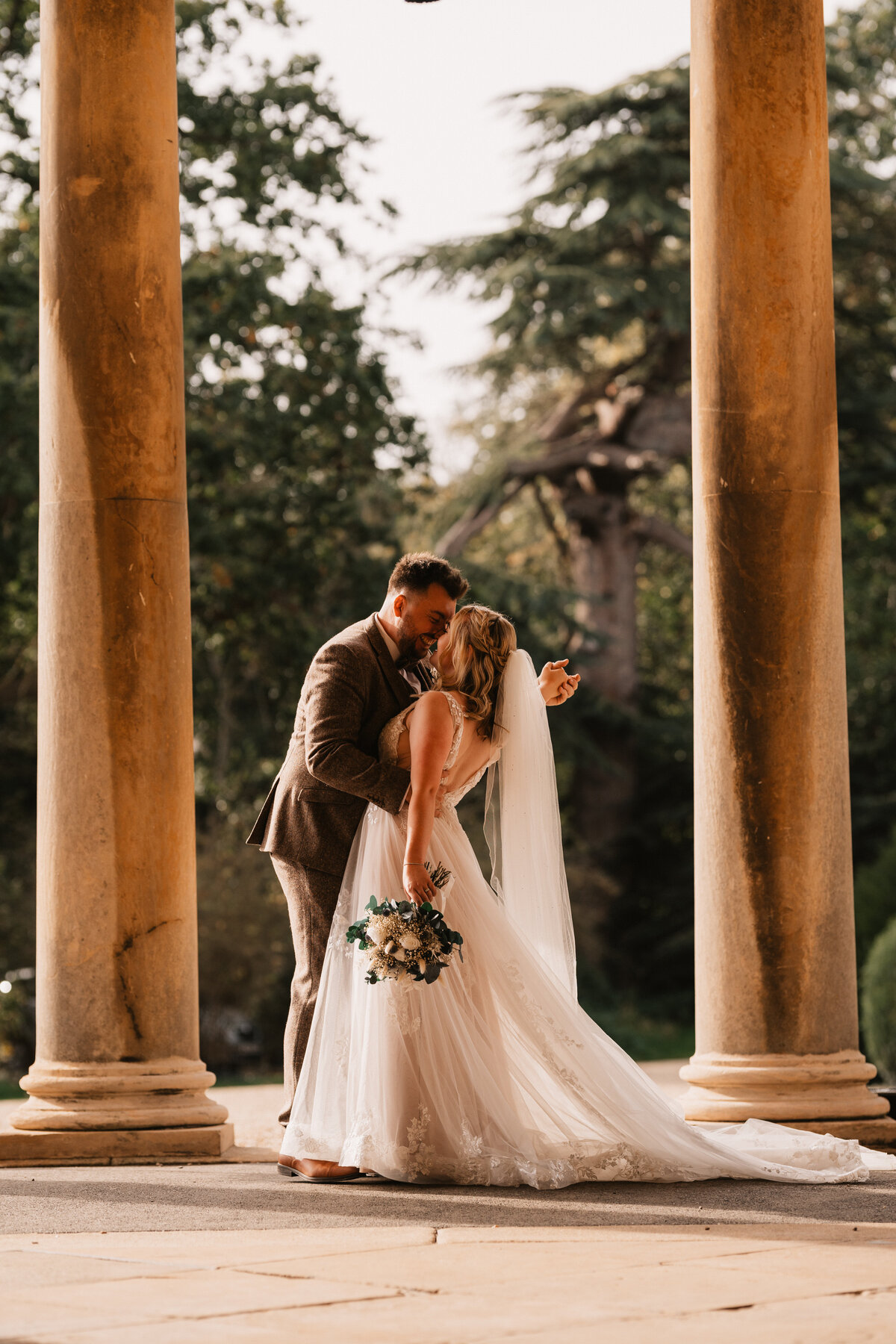 Bride and groom sharing a romantic moment under stone columns at Prestwold Hall in Loughborough, surrounded by elegant architecture and golden light.