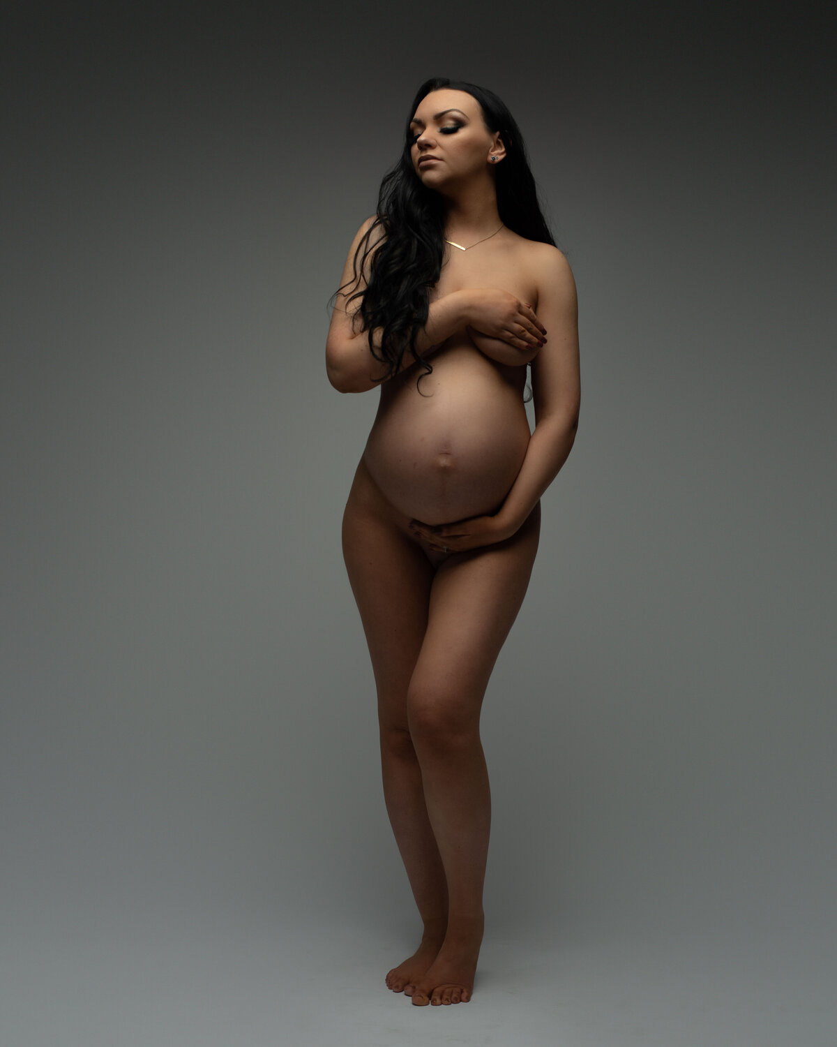 Nude maternity photograph with long haired woman standing holding one hand on tummy and other hand covering breasts.