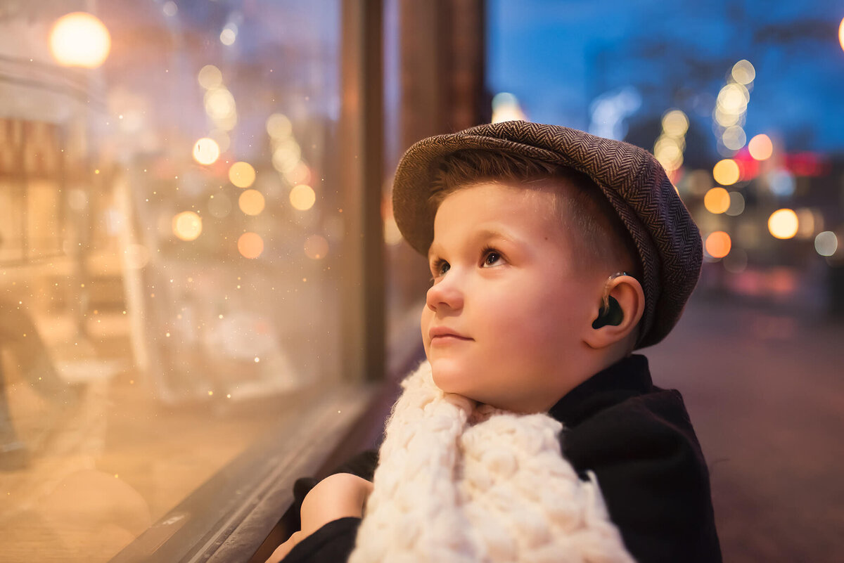 A little boy wearing a hat looks into a storefront window that is glowing. Winter portrait taken by Brittany Danielle