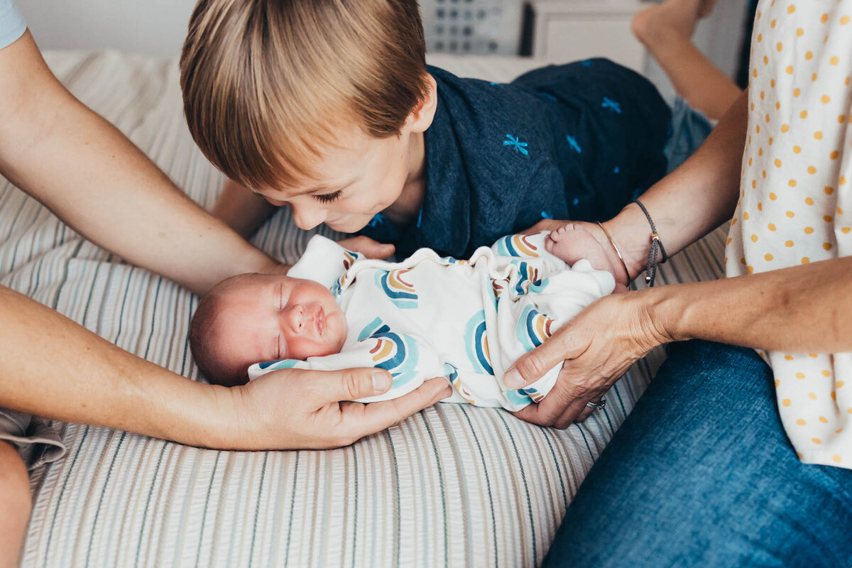 A baby lays on a bed with his parents hands on him while his big brother looks over at his adorable baby face