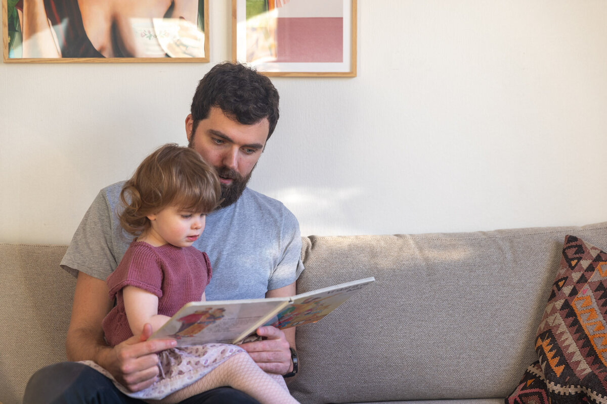 Livsstilsfoto av far som sitter i grå sofa med datter på fanget og leser bok med henne.
