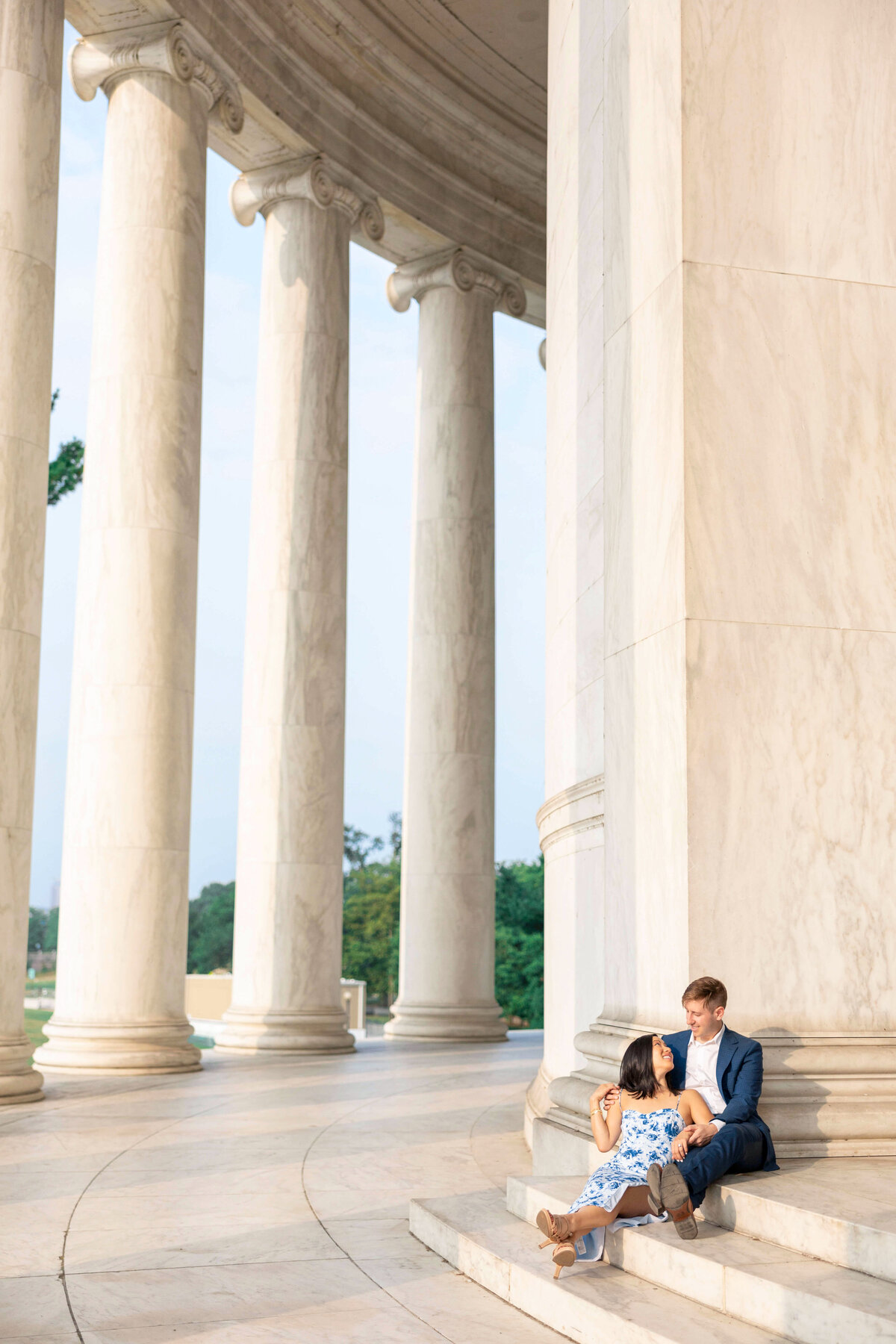 A couple sits on the steps of a grand, columned building. The woman in a blue and white dress leans against a column, while the man in a dark suit sits beside her, looking at each other affectionately. Lush greenery is visible in the background.