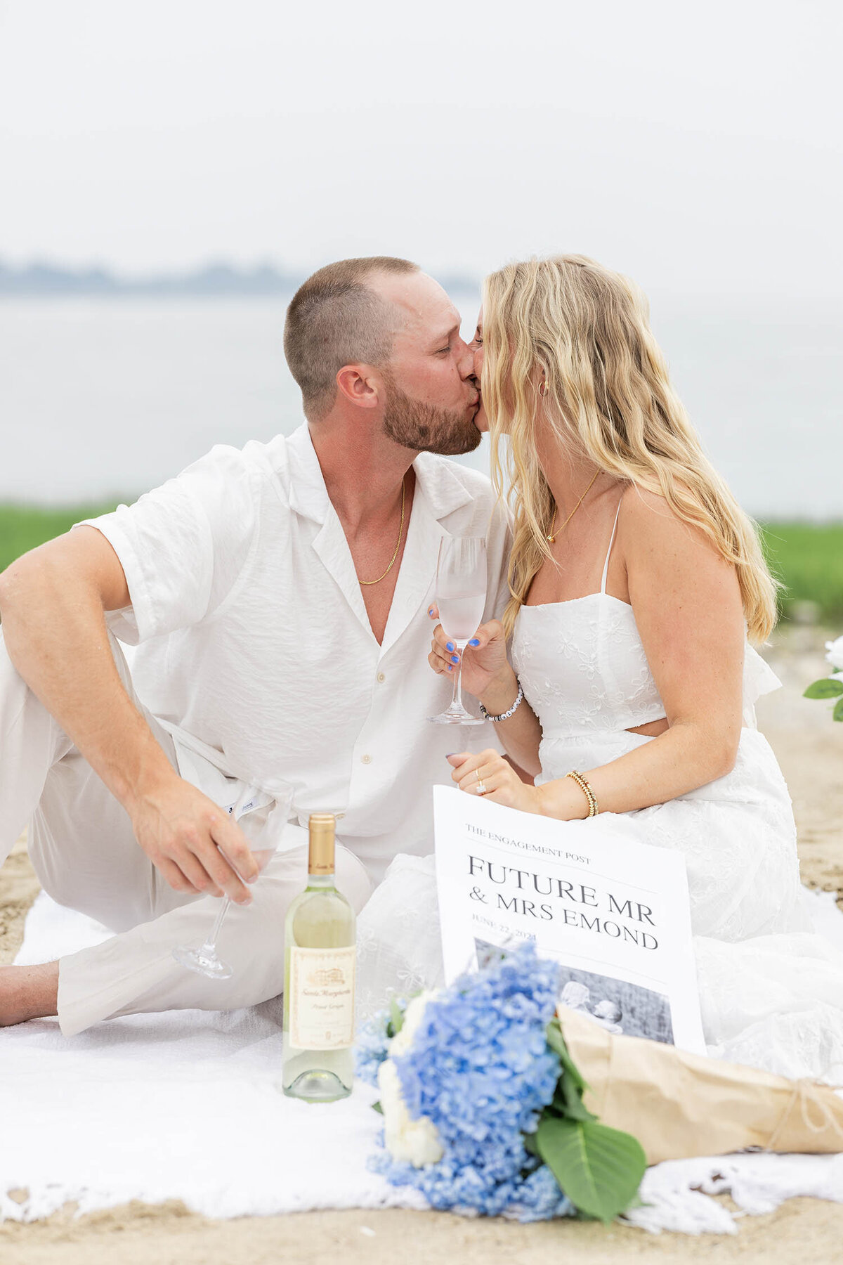 A man and woman sitting down on a blanket, shares a kiss while holding wine glasses during a coastal Connecticut session