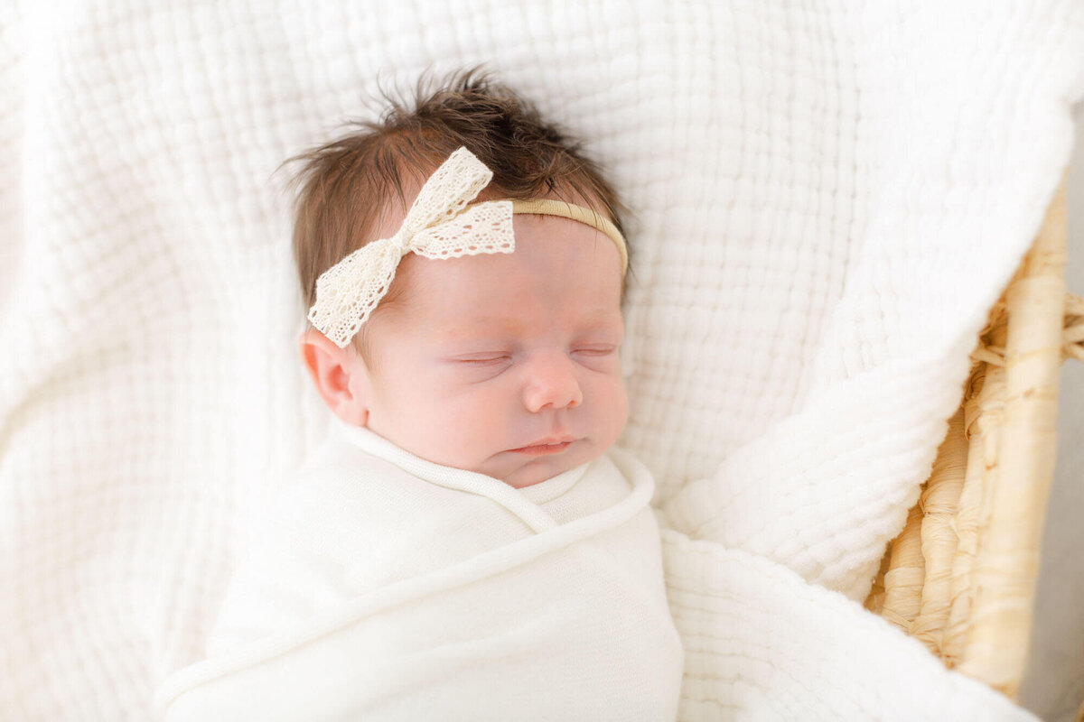 sweet baby girl with cream lace bow on her head and wrapped in a white cloth is sleeping on a textured white blanket in a Moses Basket