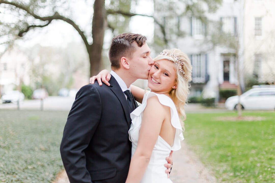 Groom kisses his brides cheek while standing on a path in the park after they eloped.