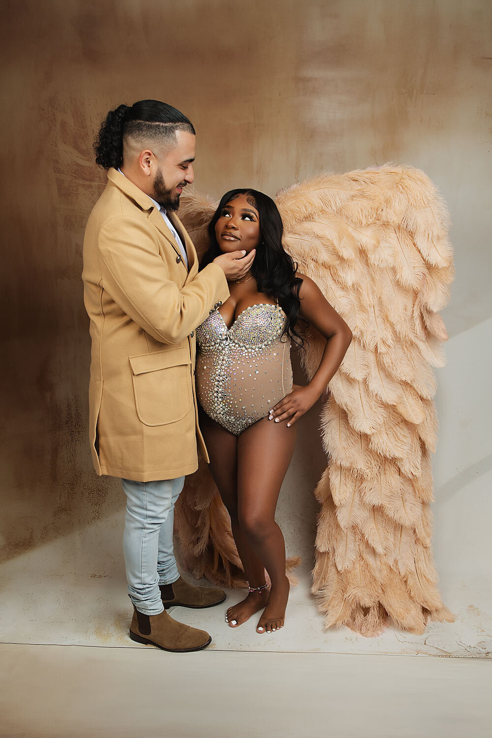 A man in a tan overcoat holds his pregnant wife's chin as she stands in a studio wearing large feather angel wings