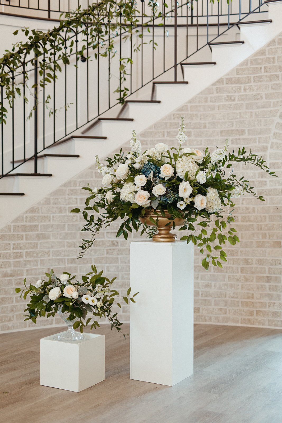 White floral arrangements on white stands in a bright and airy room with whitewashed brick staircase behind.