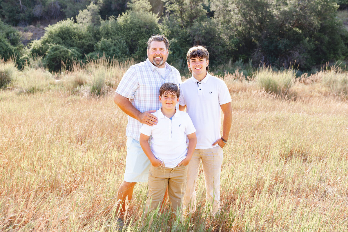 A father stands with his two sons in a tall grassy field, all dressed in coordinating white and beige outfits. The natural Midway, Utah landscape surrounds them.