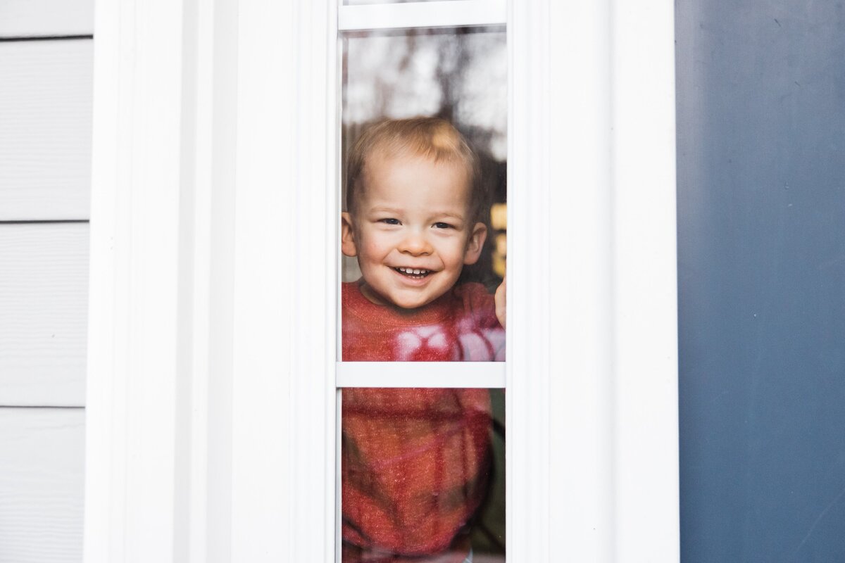 Toddler smiling behind a glass door with white frames, captured by a skilled Pittsburgh family photographer.