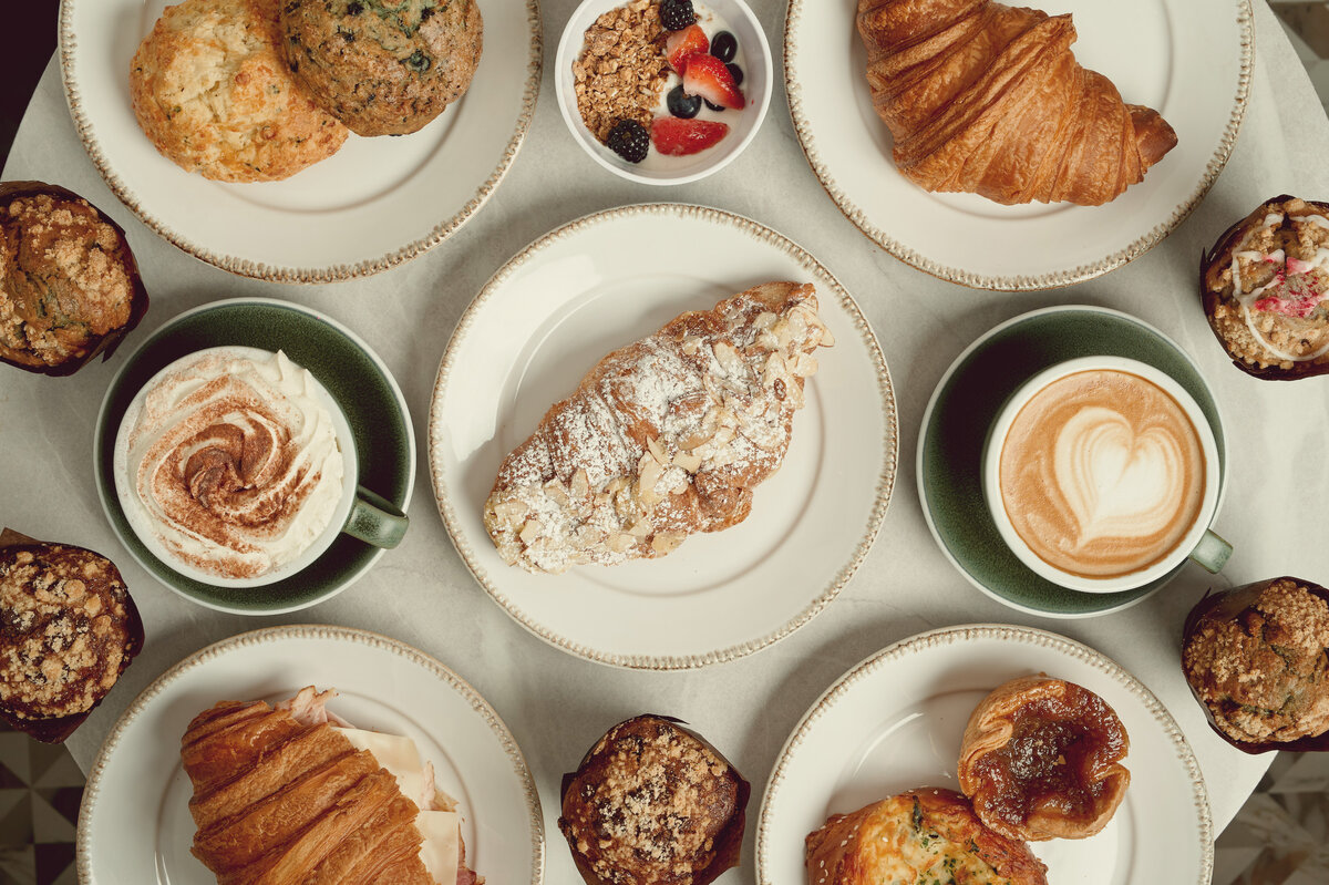 Assorted pastries, muffins, and coffee cups on a table, including croissants, muffins, granola with fruit, and latte art, representing Lyla's House specialty coffee and wine bar in Georgetown, ON.