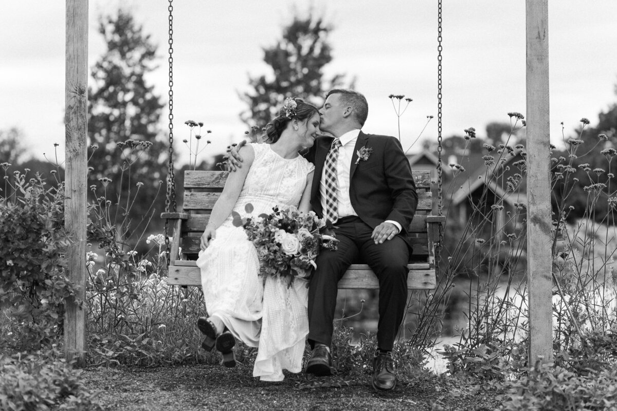 Bride-and-groom-sit-on-outdoor-swing-by-pond-and-kiss-at-Monroe-WA-wedding-venue-Pine-Creek-Nursery-photo-by-Joanna-Monger-Photography