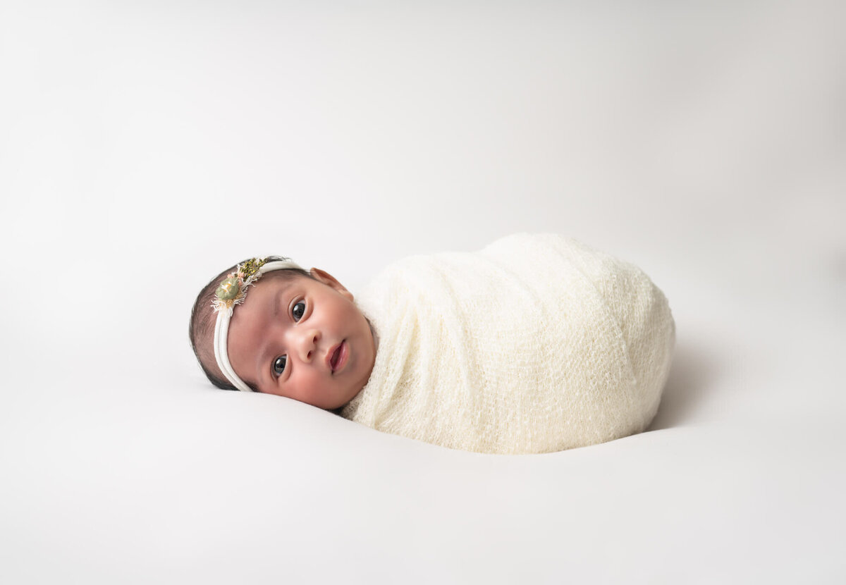 A newborn baby girl wrapped in a white blanket with a white headband stares at the camera during her portrait session with an Asheville Newborn Photographer