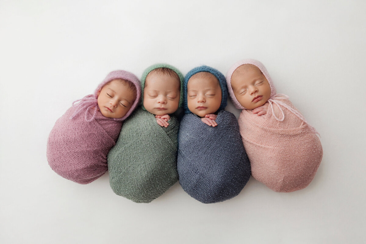 Four newborn babies are peacefully sleeping, each swaddled in a cozy knit blanket of different colors: lavender, green, blue, and pink. They are arranged side by side on a white background, creating a serene and heartwarming scene. Each baby wears a matching bonnet, adding to the adorable and uniform look.