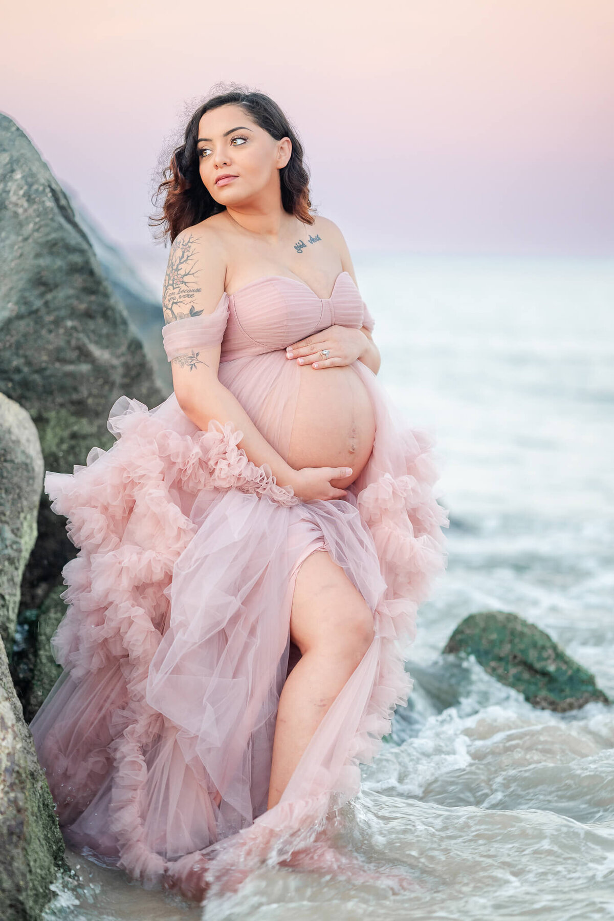 A pregnant woman wearing a pink tulle dress cradles her belly while standing in the waves at the beach.