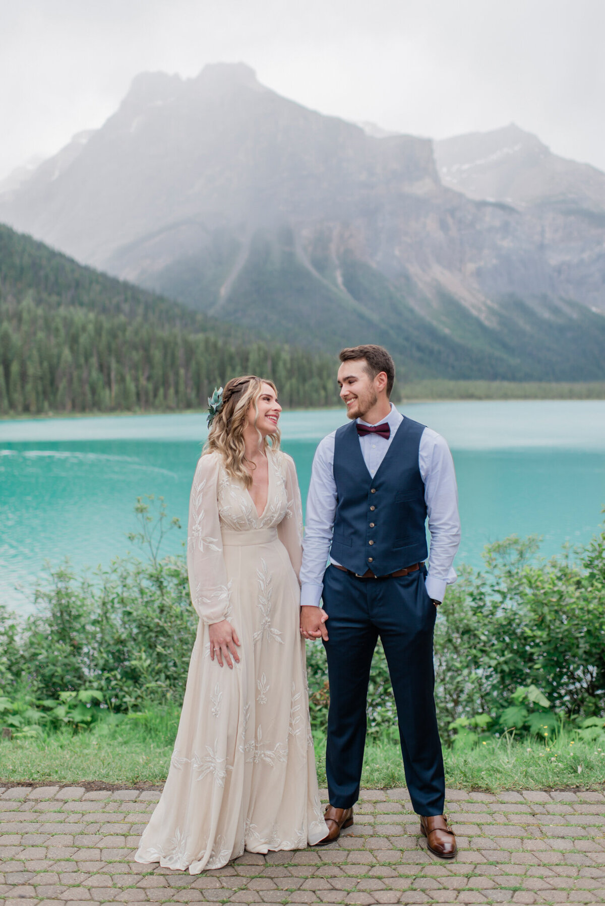 Stunning and elegant couple standing in mountains of Emerald Lake  , bride wearing gorgeous BHLDN cream gown, groom in navy blue suit vest, captured by Kaity Body Photography, elegant film inspired wedding photographer in Calgary, Alberta. Featured on the Bronte Bride Vendor Guide.