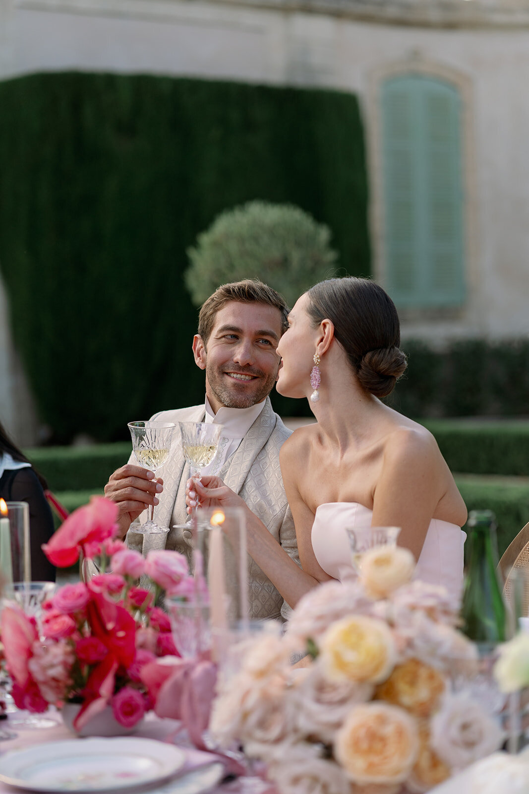 Bride and groom smiling and toasting at outdoor dinner table decorated with pink floral arrangements at Château de Tourreau.