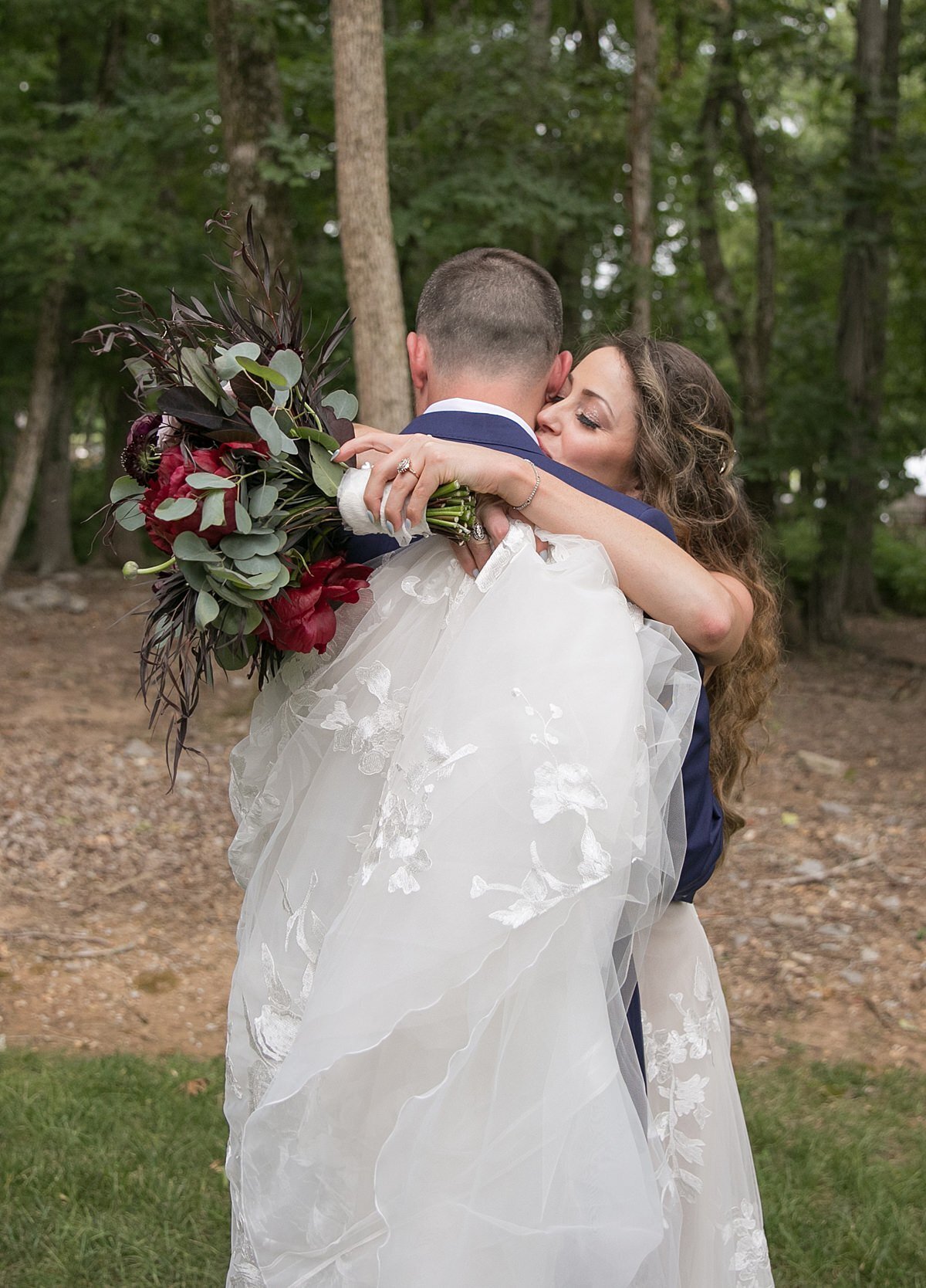 The bride and groom, wrapped up in the lace train of her wedding gown, embrace and kiss in the woods while holding the bridal bouquet of burgundy ranunculus, blush roses, red peonies and burgundy roses.
