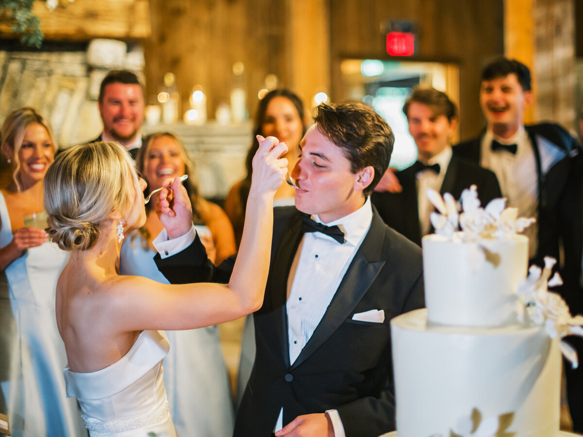 Bride and groom share a playful cake cutting moment at their wedding reception at Old Edwards Inn in Highlands, North Carolina, surrounded by cheering wedding party.