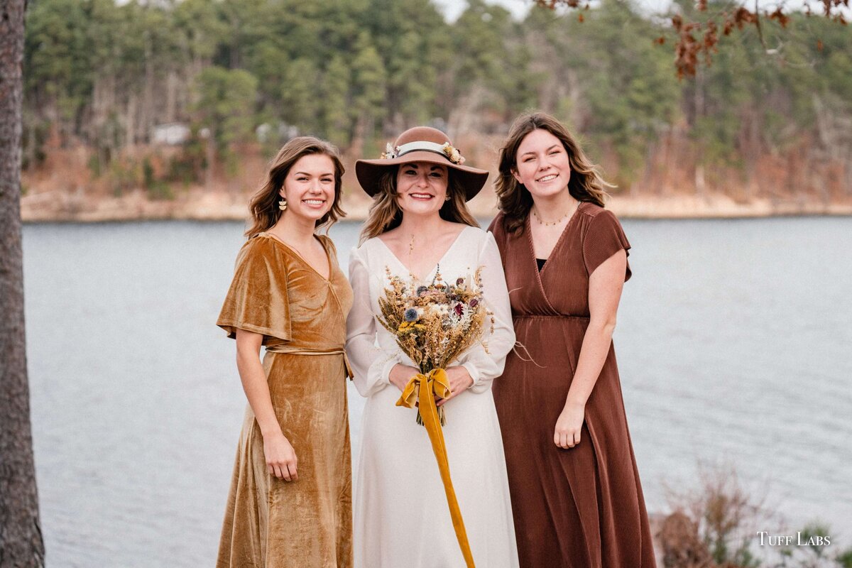 Bride holds her dried floral bouquet and stands by bridesmaids with water in the background.