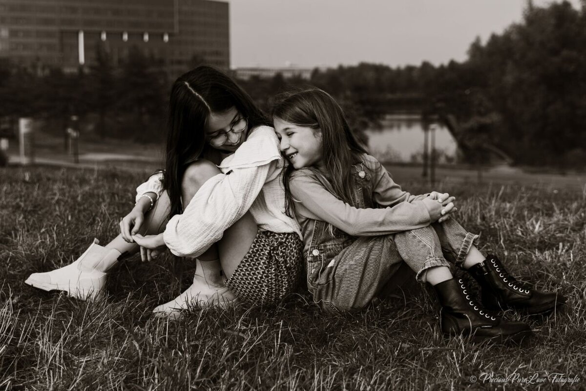 Black and white portrait of two sisters sitting in grass, leaning in and sharing a giggle.