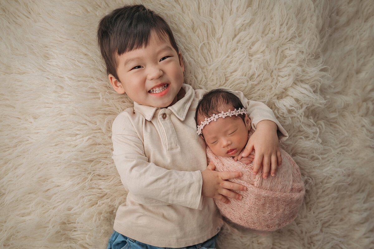 smiling new big  brother  laying down on his back  on a white rug while holding his newborn baby sister who is wrapped in a pink swaddle and wearing a floral headband