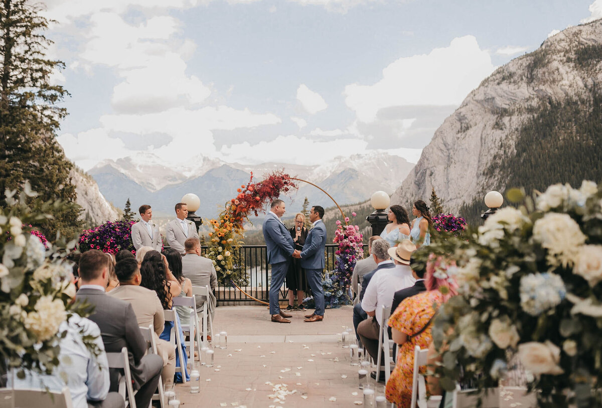 Outdoor wedding ceremony with a mountainous backdrop at the Banff Springs Hotel in Banff, Canada. A couple stands at the altar beneath a floral arch, surrounded by guests seated on both sides of the aisle, creating a modern-day fairytale wedding.