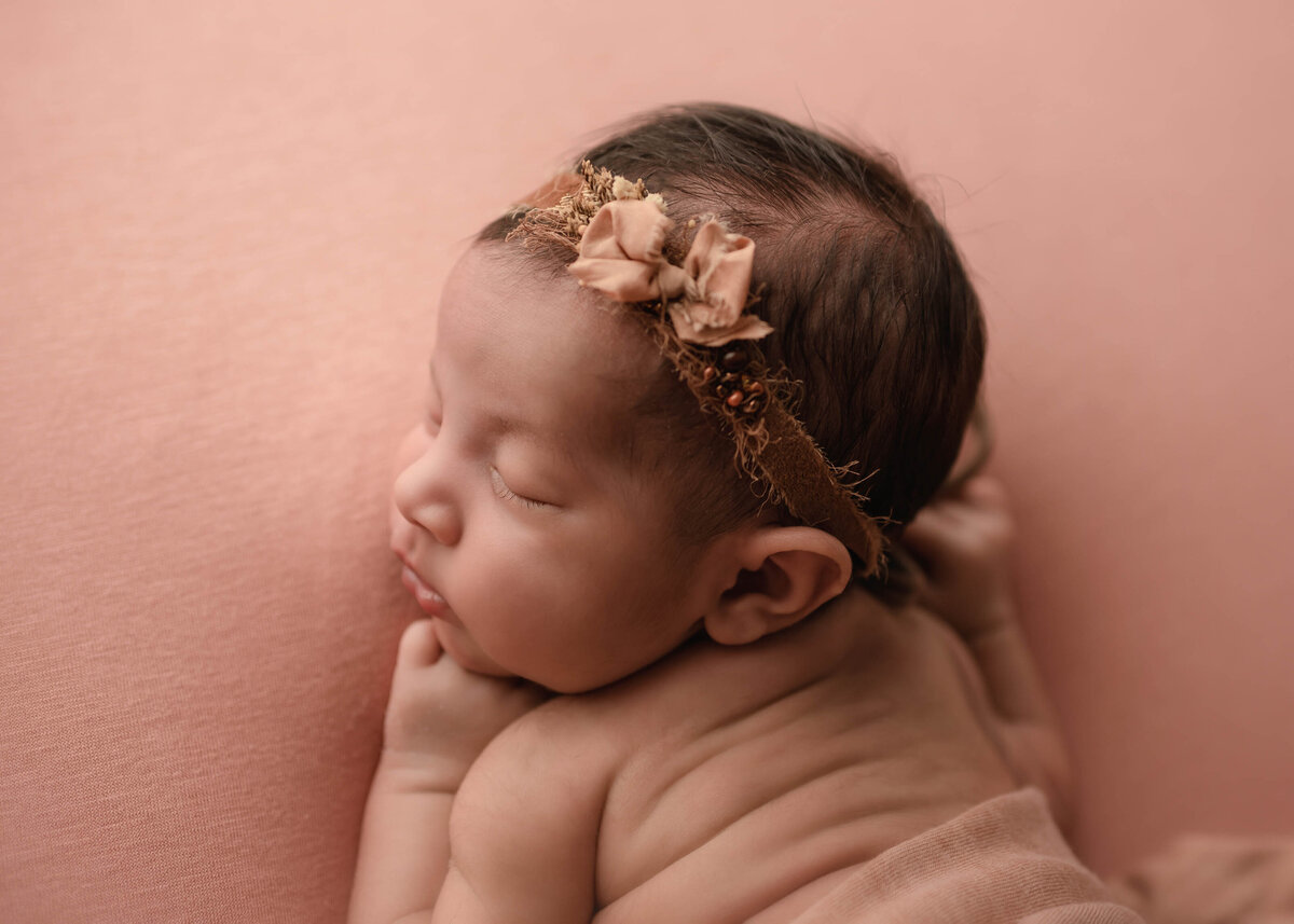 Baby sleeping on belly on rose gold knit blanket wearing matching bow and floral headband. Cute wrinkles on the baby's back.