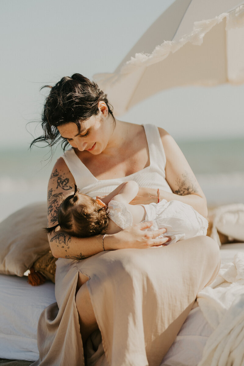 Maman en train d'allaiter sa fille, assise sur un lit et protégée par une ombrelle dans un décor de plage. Capturée par Laura, photographe maternité en vendée.