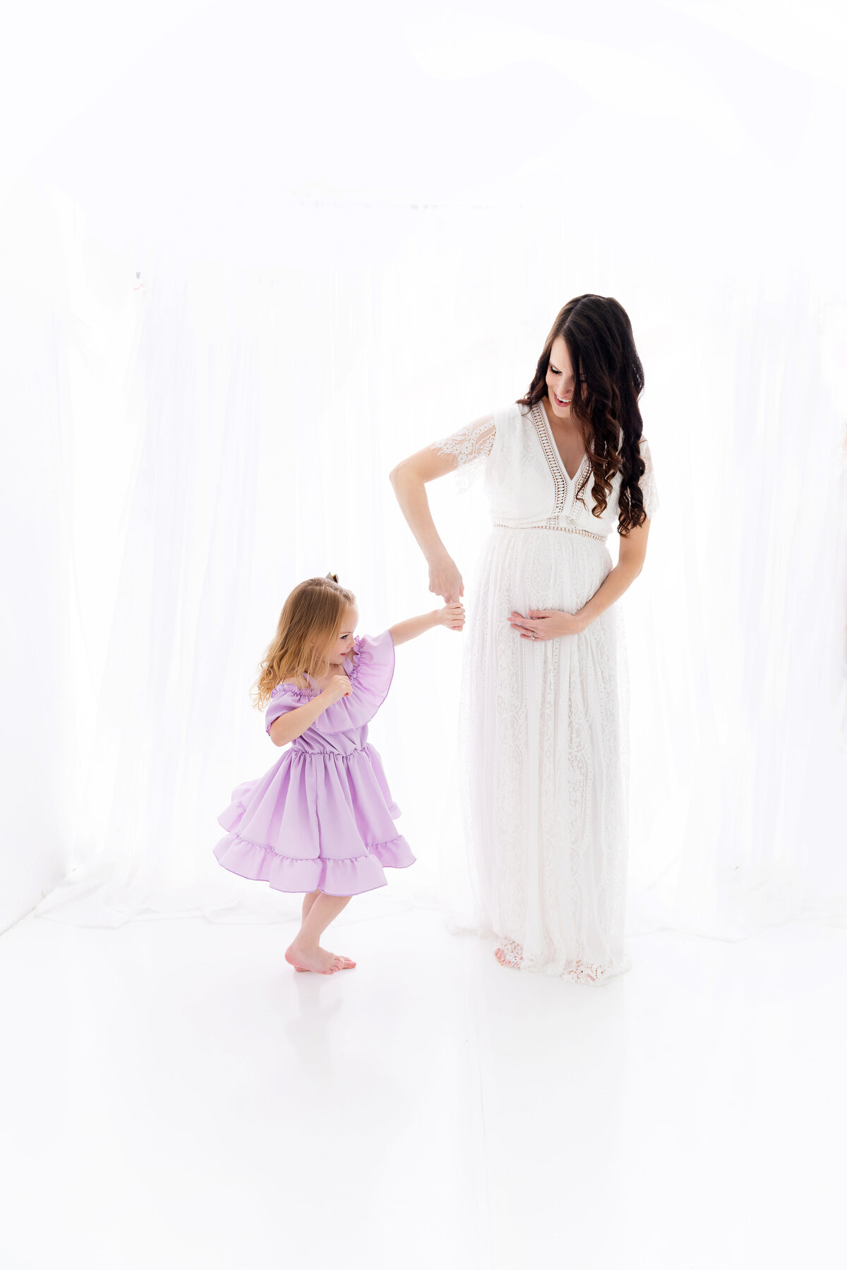 A pregnant mom spins her daughter around in a white gown for maternity photos