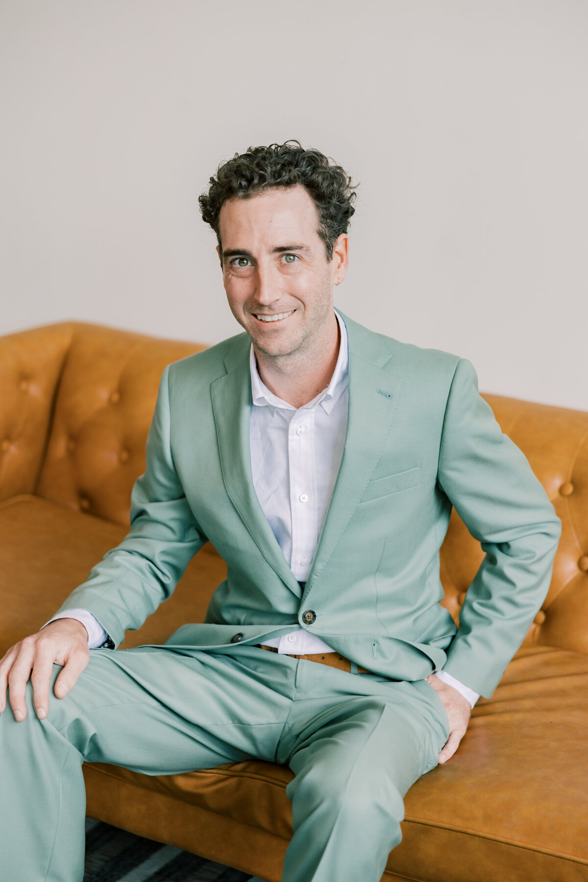 Brendan wearing sage green Indochino suit sitting on leather sofa before wedding at Fetewell in Baltimore.
