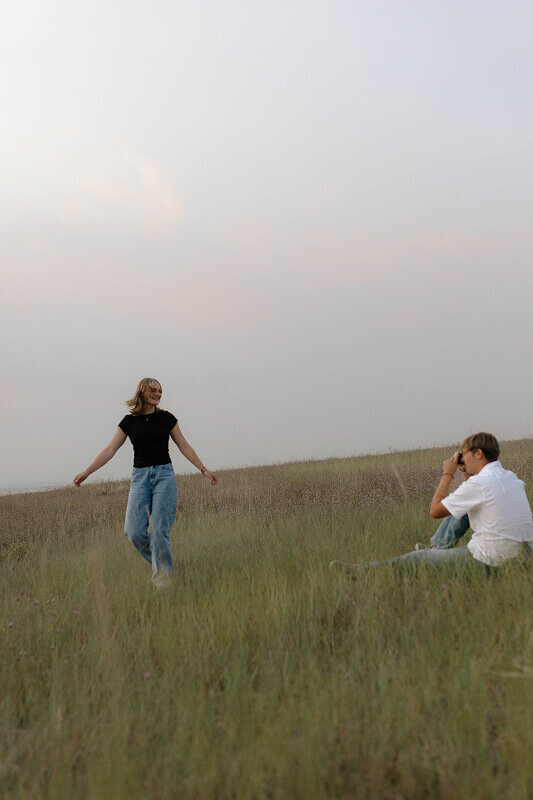 A man takes a photo of his girlfriend as she twirls in a grass field.