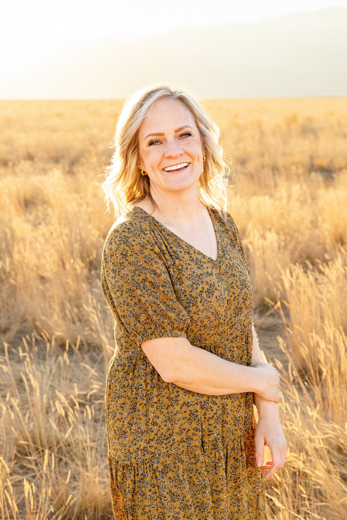 A mother in a patterned dress stands in the sunlit field, smiling with golden light illuminating her blonde hair.