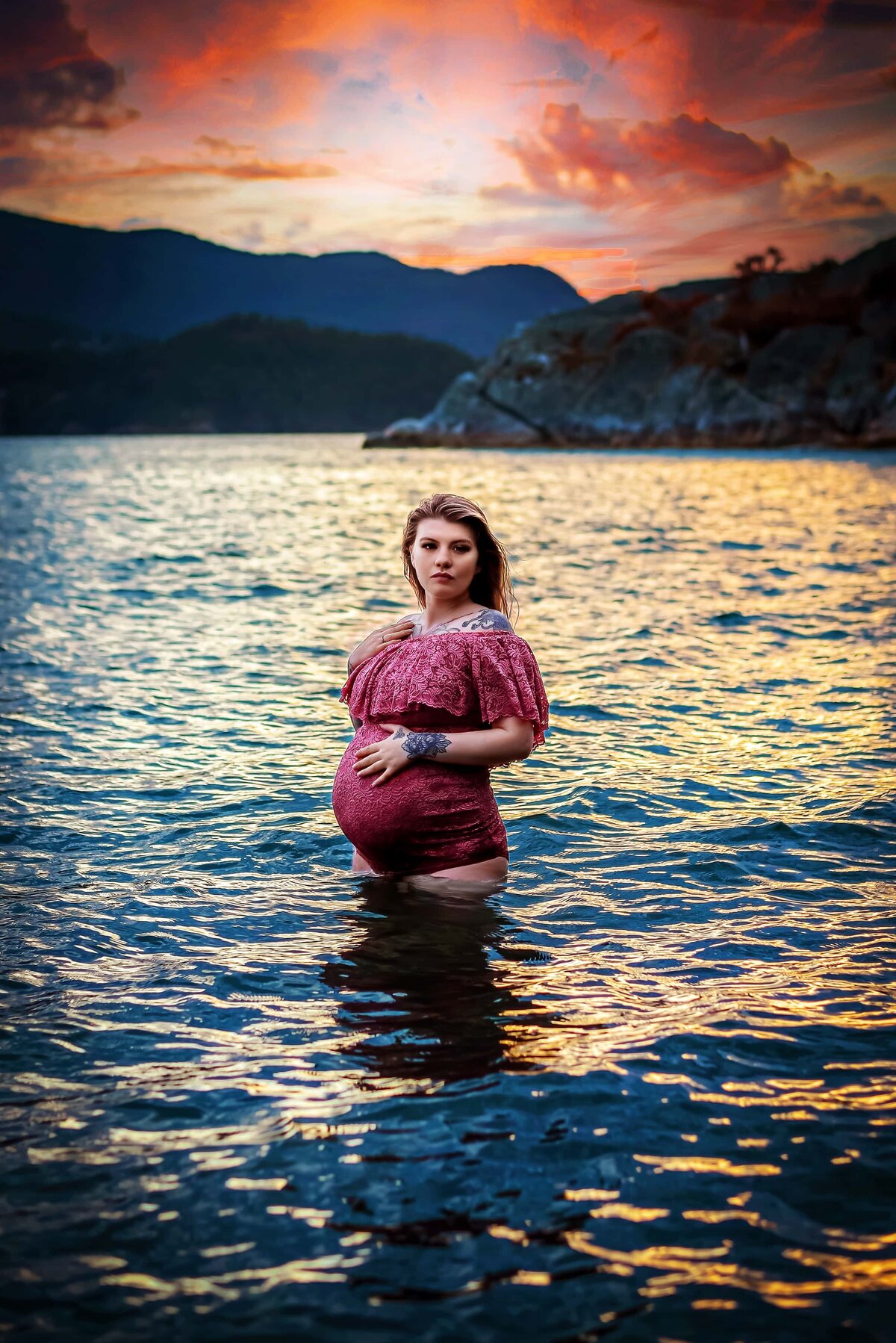 Pregnant woman in pink lace bodysuit in the water at sunset.