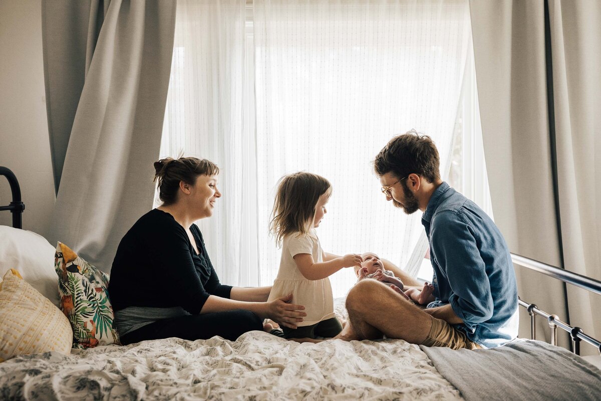 Mom, dad, and toddler girl are looking sweetly at the newborn baby in bed.
