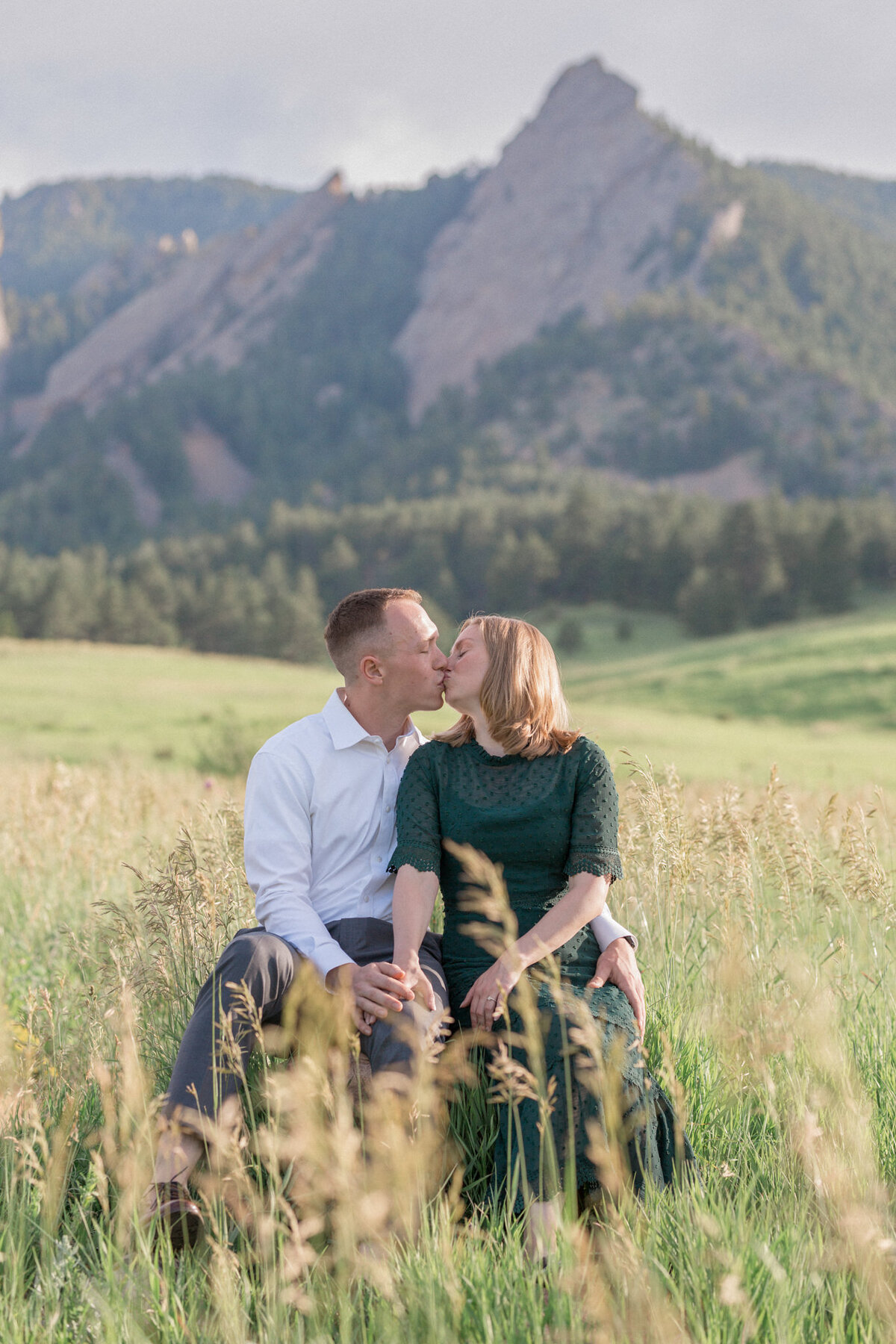An engaged couple share a kiss while seated in front of a stunning view of the Boulder Flatirons