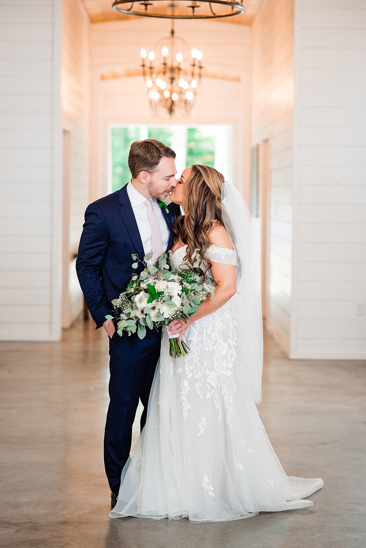 The bride and groom kiss in a large white room with a polished wood floor and a brass chandelier hanging above the open door behind them. The bride is wearing a long sheer veil and an off the shoulder wedding dress with a fitted lace bodice and full flowing sheer skirt with lace details. She is holding a large bouquet of white flowers and greenery. The groom has one arm around the bride's waist and the other hand in is pocket. The groom is wearing a navy suit with a white shirt and blush tie.