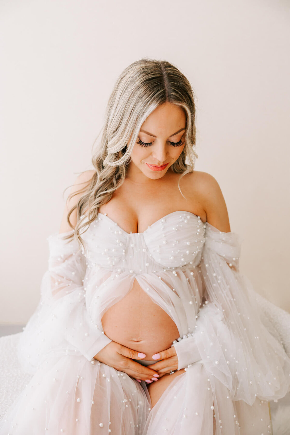 portrait of beautiful blonde woman holding pregnant belly wearing gown made of pearls and tulle in studio photography session