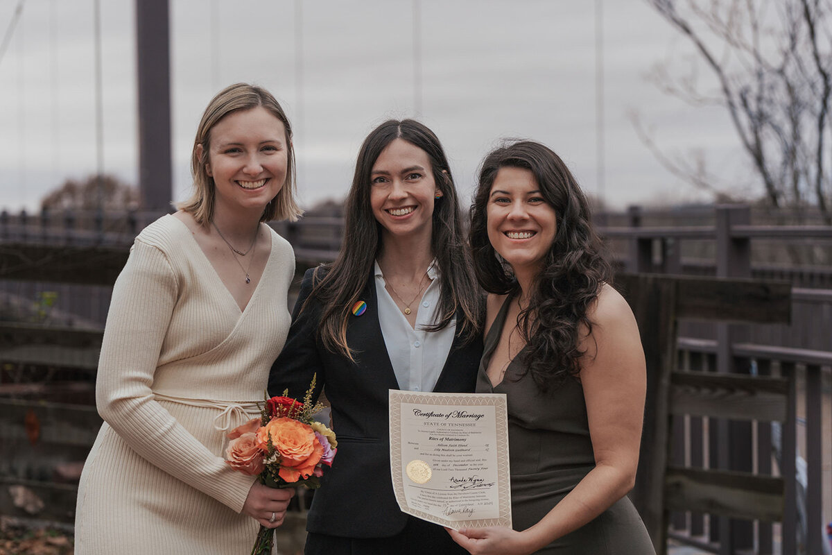 Women smile standing next to their officiant with their marriage license in hand 