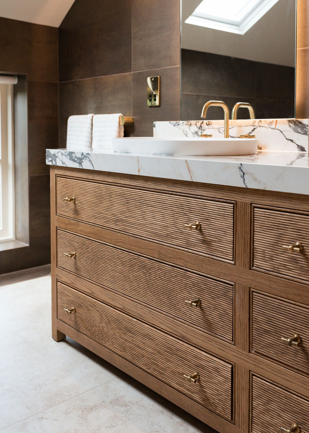 A modern bathroom, featuring a double sink vanity with marble countertops and wooden cabinets.The room is finished with dark tiled walls and light tiled floors.