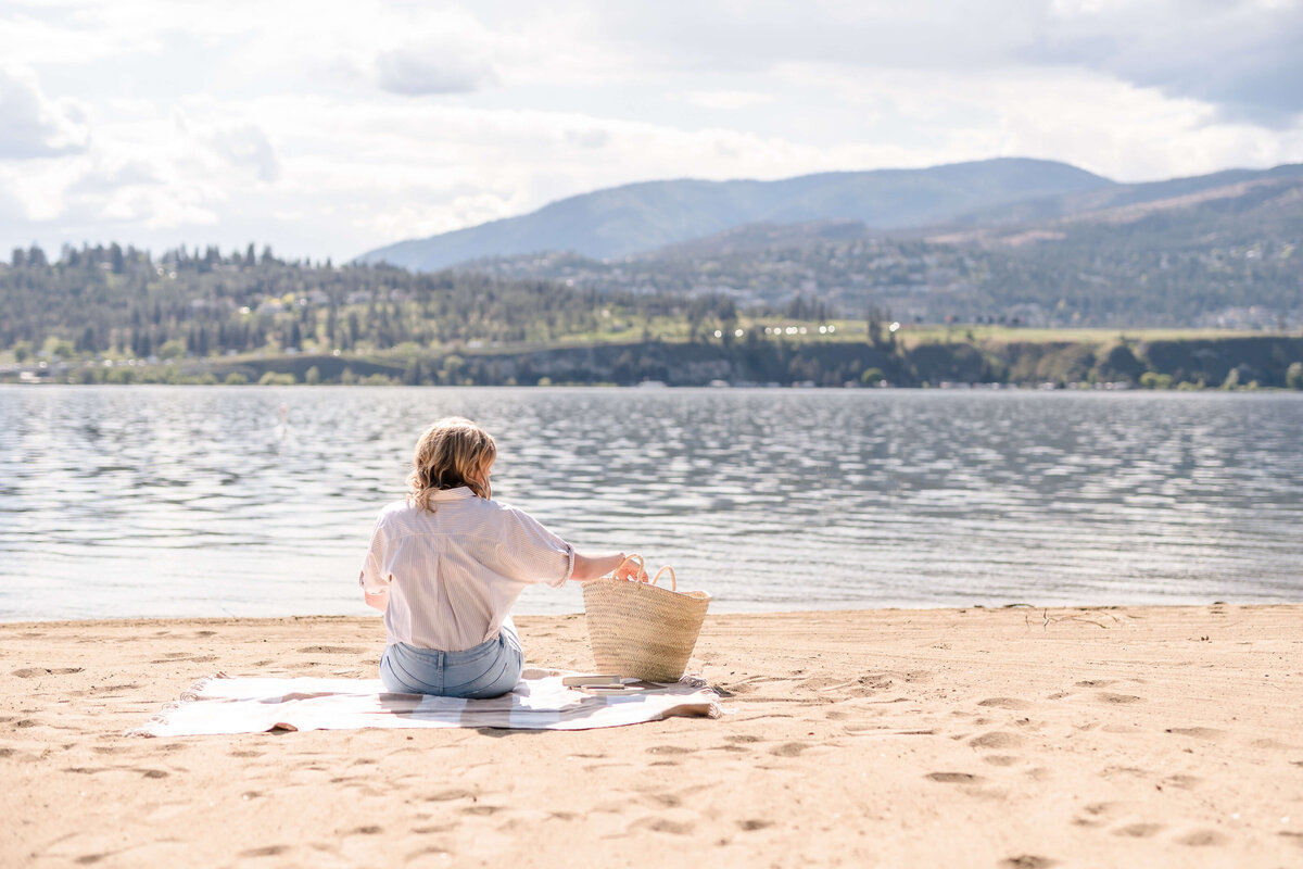 Woman sitting on blanket at Okanagan Lake beach reaching into woven tote on sunny day.