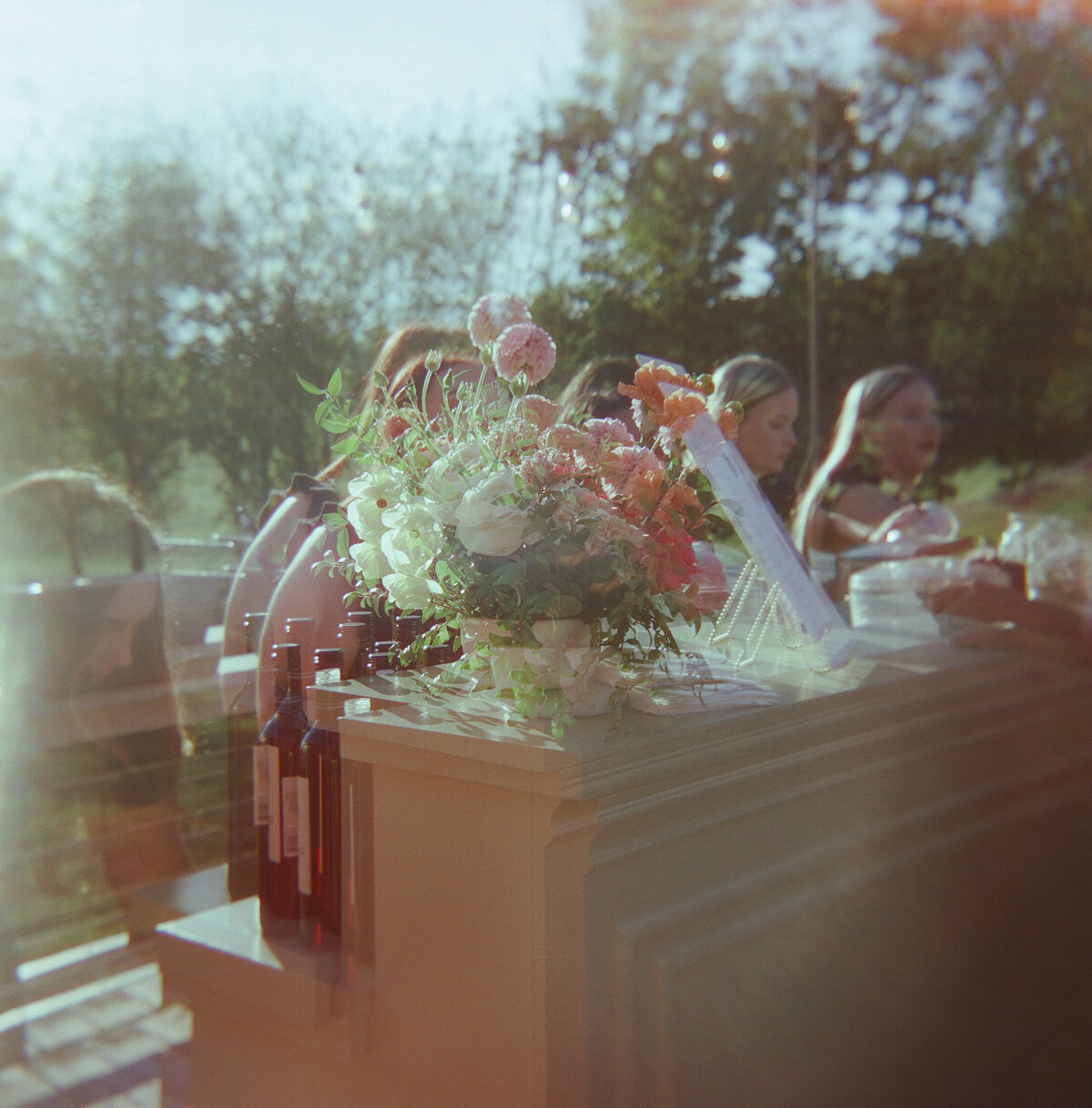 Dreamy film photograph of a lush wedding floral arrangement with coral, pink, and white blooms on an outdoor bar, capturing warm natural light and organic garden-style design.
