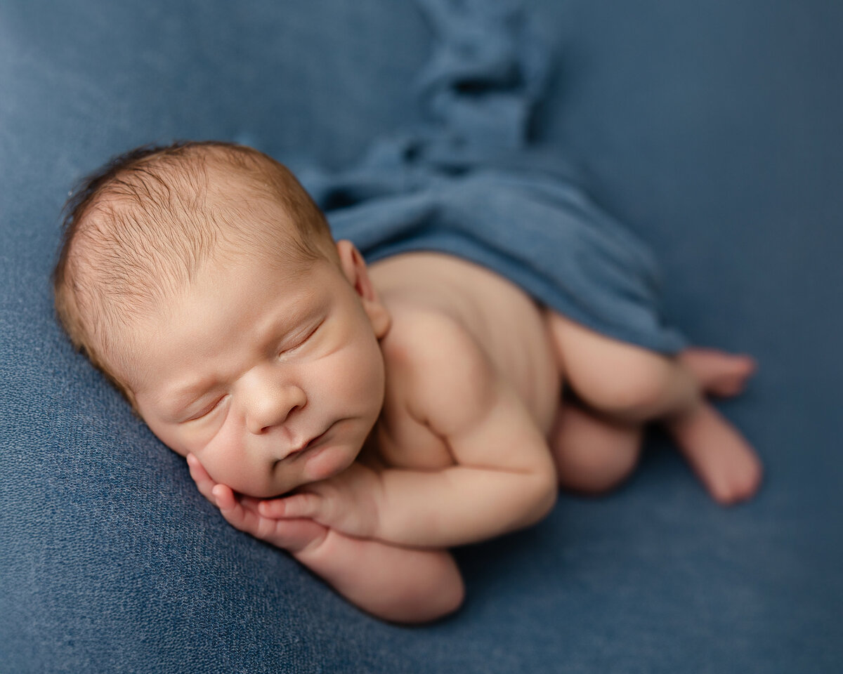 Newborn baby boy peacefully posed on a soft blue backdrop, with a matching wrap gently draped across him. Captured by Inland Empire newborn photographer Kaitlyn Dawn Photography.