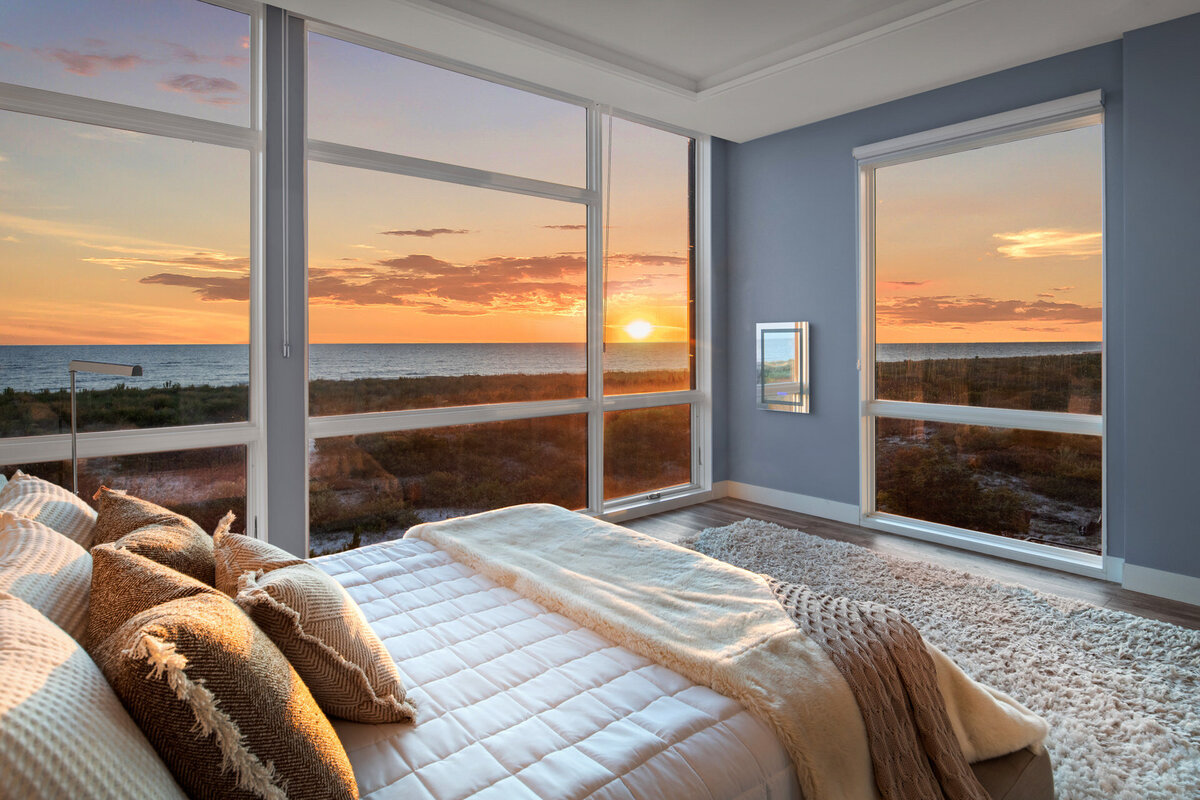 A bedroom with large windows showcases a stunning ocean sunrise view, expertly photographed by Svetlana Leahy of Refined Capture, based in Ocean City, Maryland. The room features a neatly made bed with pillows and a beige throw, light blue walls, and a plush white rug on the floor.