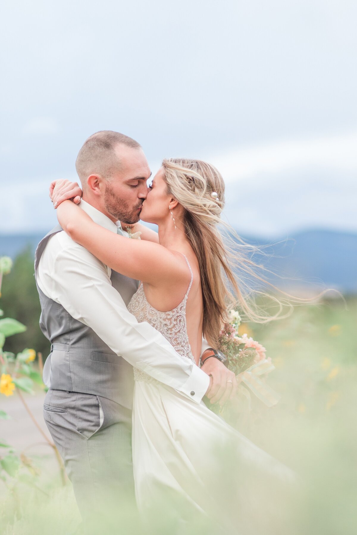 Bride and groom kiss in a field of tall grasses with mountains in the background